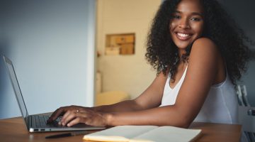 People, lifestyle, modern technology, job and communication concept. Attractive young dark skinned female freelancer with curly hair having pleased look, enjoying distant work, using generic laptop